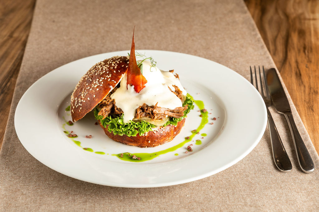 Gurm&aacute;nsk&yacute; burger s trhan&yacute;m masem, sal&aacute;tem a smetanovou om&aacute;&ccaron;kou v sezamov&eacute; bulce, elegantn&ecaron; naserv&iacute;rovan&yacute; na b&iacute;l&eacute;m tal&iacute;&rcaron;i